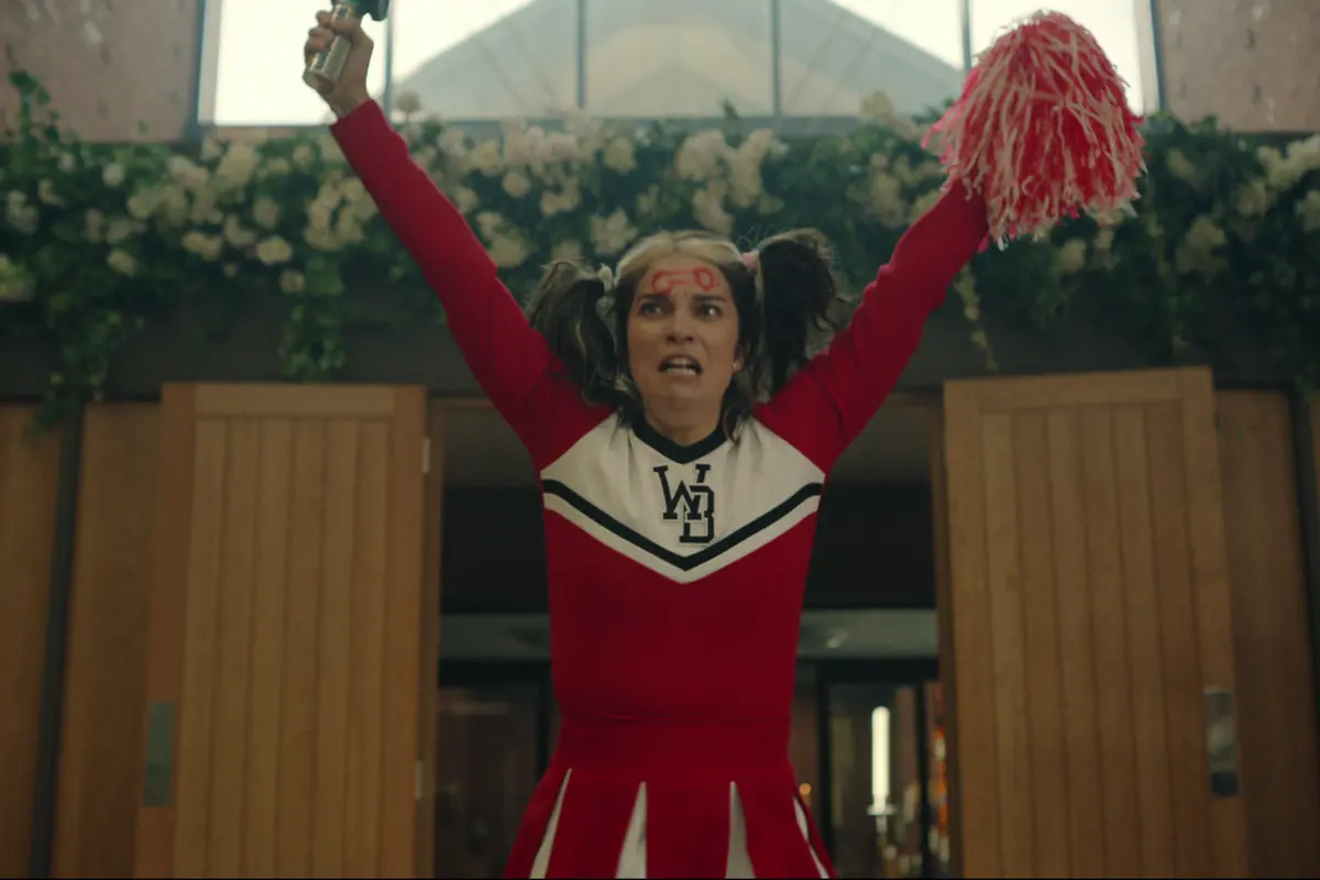 Mulher jovem com cabelo preso, veste blusão e saia, segurando pompons de líder de torcida nas mãos.