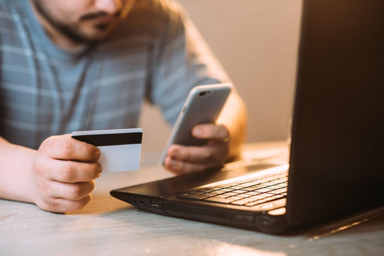 homem segurando cartão e celular diante de notebook