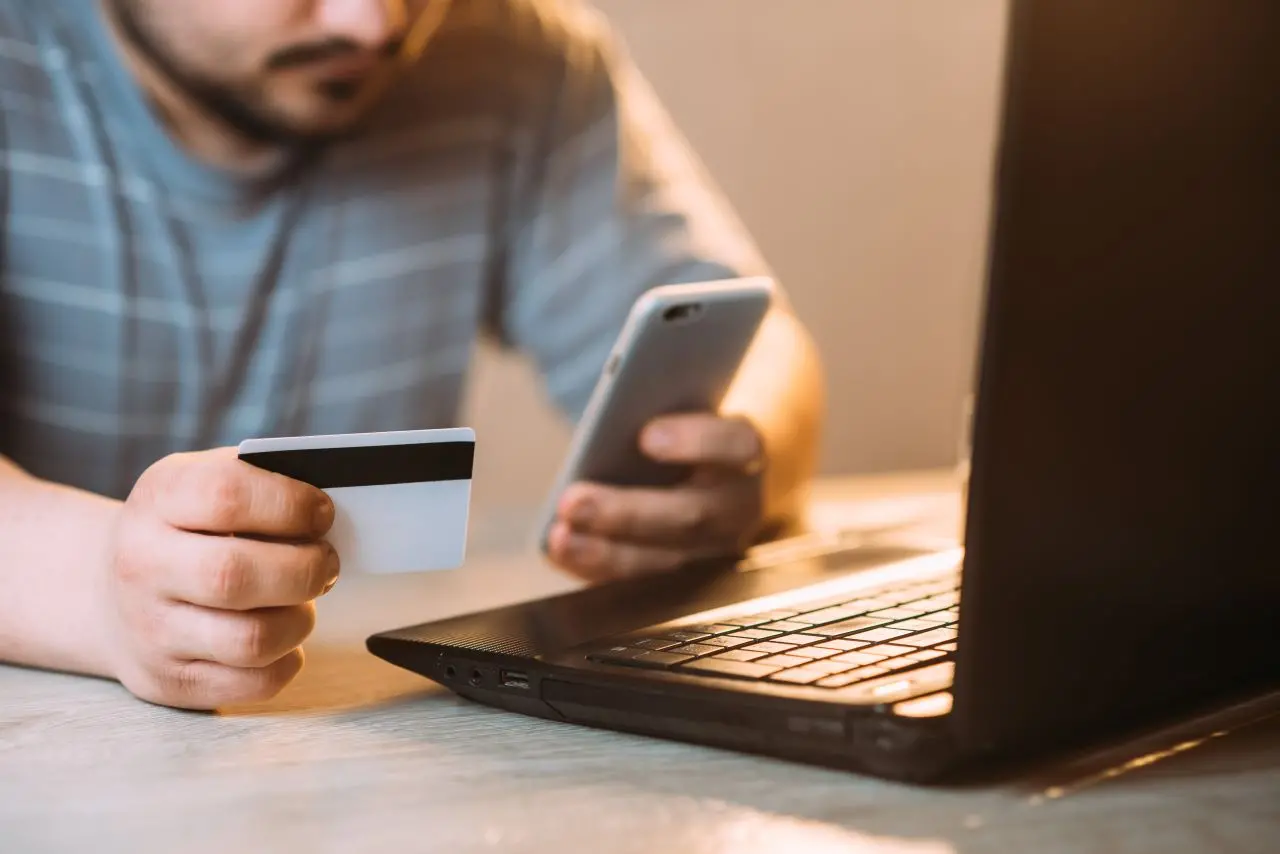 homem segurando cartão e celular diante de notebook