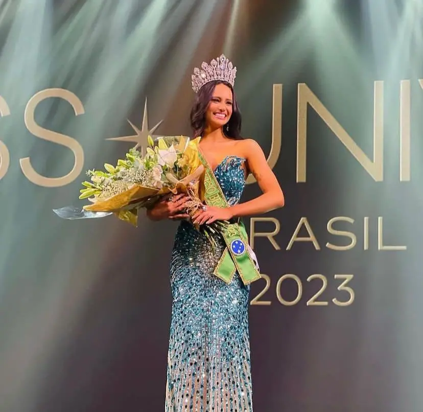 Maria é uma mulher branca, alta, de cabelos pretos e ondulados. Na foto, está usando uma coroa, um vestido azul e segurando um buquê