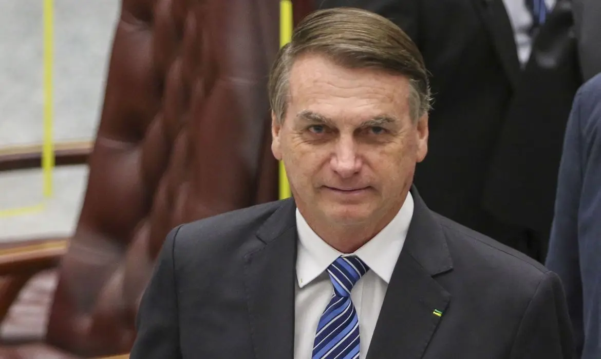 Bolsonaro é um homem branco, idoso, e tem cabelo bem penteado para o lado. Na foto, está de terno, camisa branca e gravata azul listrada