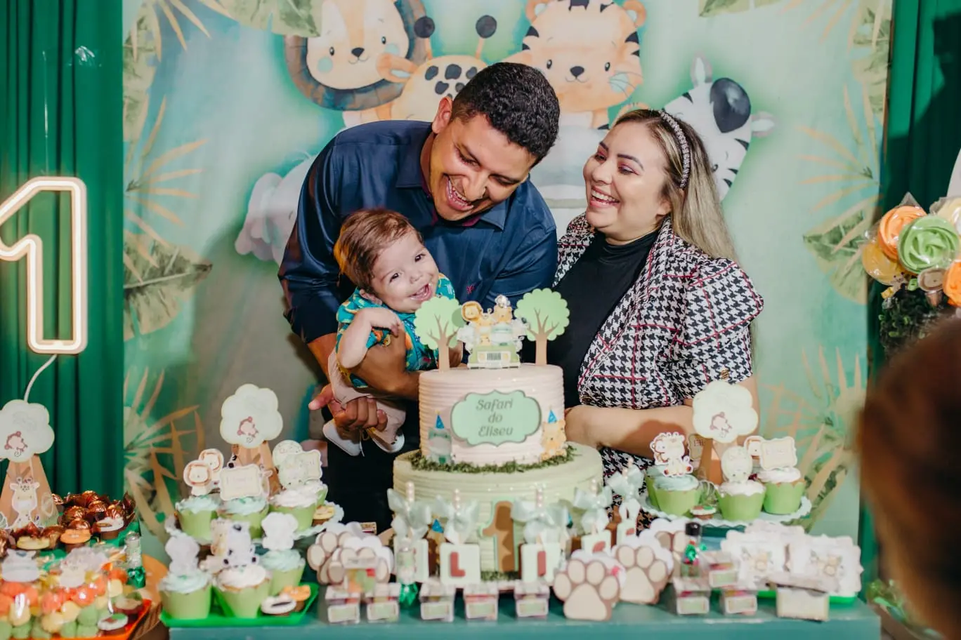 Aniversário de 1 ano do Eliseu. Pai, mãe e criança atrás de bolo e mesa decorada