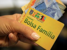 Mão com unhas grandes e pintadas segura cartão do Bolsa Família e cédulas de 100 e 20 reais