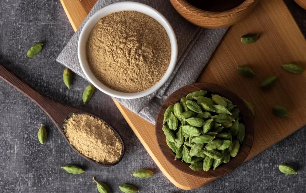 Pó de cardamomo dentro de um recipiente branco ao lado de uma colher de pau