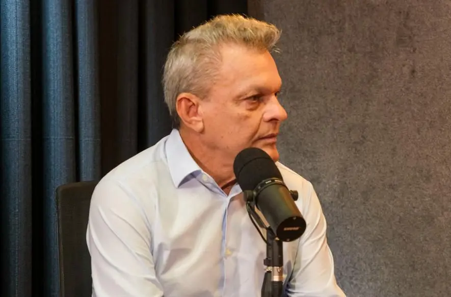 José Sarto é um homem branco e de cabelos brancos. Ele está falando ao microfone em um estúdio de podcast