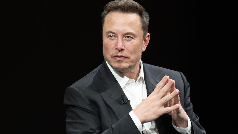 Elon Musk é um homem branco e de cabelo preto penteado com topete. Na foto, ele está de terno preto e camisa branca por baixo