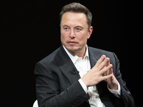 Elon Musk é um homem branco e de cabelo preto penteado com topete. Na foto, ele está de terno preto e camisa branca por baixo