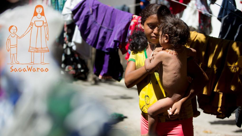 Mulher indígena venezuela cabelos cacheados segurando uma criança nos braços em meio a um varal com roupas coloridas