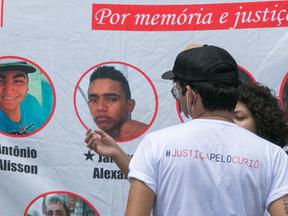 Pessoas com camisa pedindo justiça pelo Curió em frente a faixa com fotos das vítimas