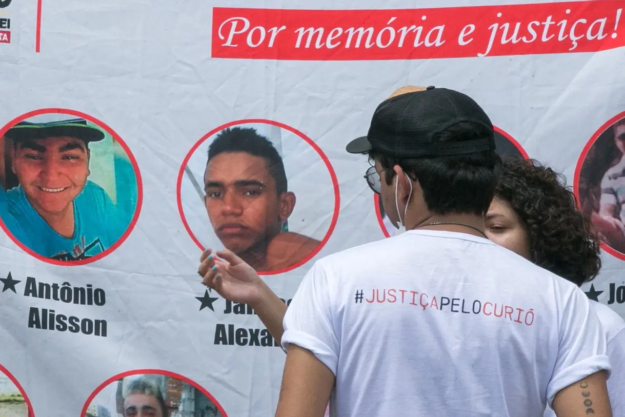 Pessoas com camisa pedindo justiça pelo Curió em frente a faixa com fotos das vítimas