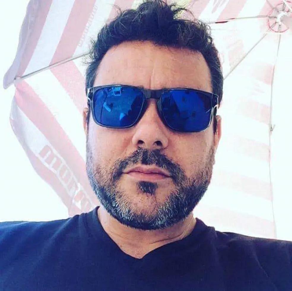 Amândio é um homem branco, de barba e cabelo preto. Na foto, ele está de blusa preta e óculos espelhados