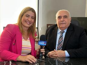 A apresentadora Natália Varela, mulher branca e loira, de blusa rosa, segura um microfone. Ao lado dela, um homem branco, de terno preto é entrevistado.