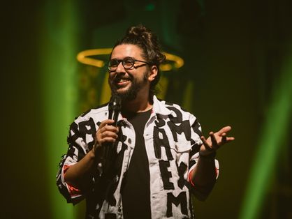 Humorista Moisés Loureiro no palco, com figurino do espetáculo Deixemos de Coisa; camisa branca estampada com letreiro preto