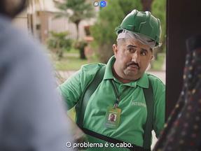 Humorista vestido de técnico de internet chega a uma casa. Ele usa roupas e capacete verde e um bigode.