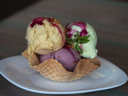 A foto contém a imagem de um sorvete de três bolas e cobertura em uma casquinha apoiados em cima de um prato