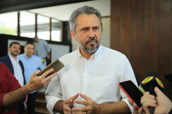Elmano de Freitas usando uma blusa social branca, com microfones ao redor enquanto concede entrevista.