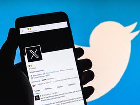Mão segurando celular mostrando perfil da rede social 'X', antigo Twitter