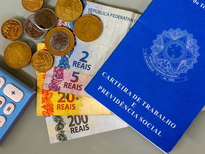 Imagem mostra carteira de trabalho com notas e moedas de real, e calculadora