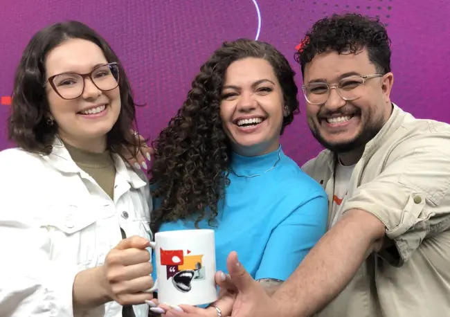 Três apresentadores do Podwyden sorriem para a foto e seguram uma caneca.