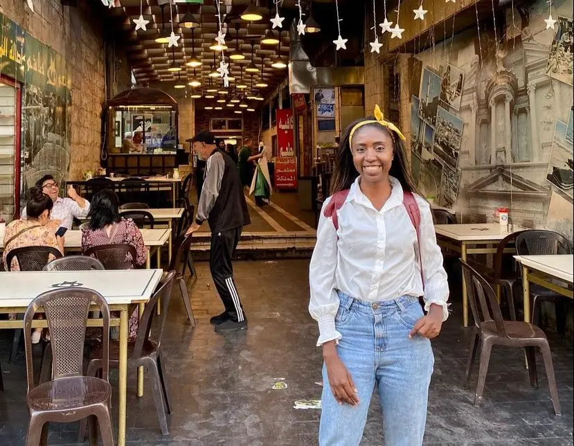 Nataly é uma mulher negra de longos cabelos castanhos. Ela veste camisa branca e calça jeans. Ela está na frente de um restaurante e de mochila