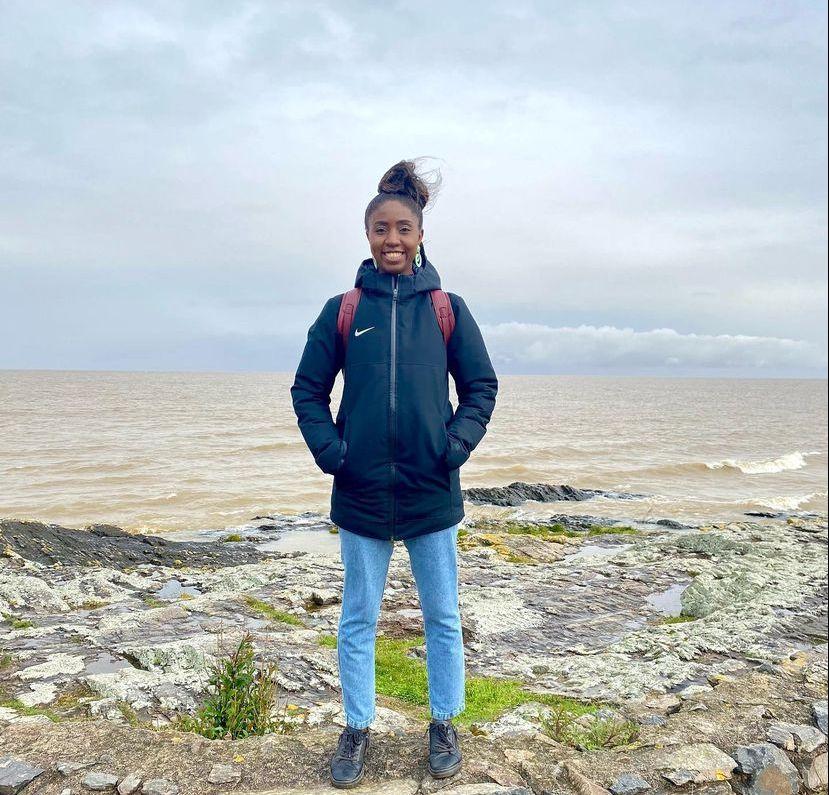 Nataly é uma jovem negra e na foto ela está com casaco azul escuro e calça jeans clara no topo de uma paisagem