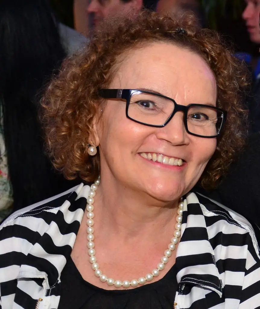 Liduína é uma mulher de meia idade, de cabelos cacheados curtos, ruivos. Na foto, ela usa óculos de aro preto, grosso, e roupa listrada em preto e branco