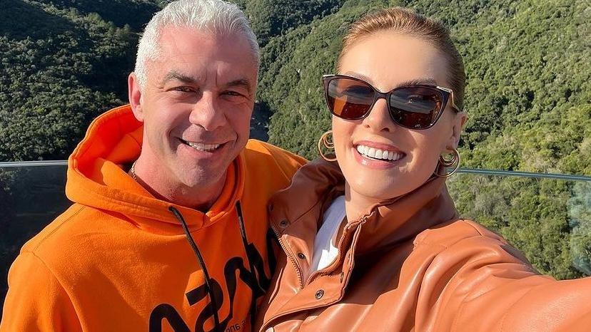 Alexandre Correa e Ana Hickmann são brancos. Ele tem cabelo raro, branco, e ela tem cabelo loiro longo. Ambos estão com casaco laranja