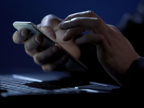 Imagem mostra mãos digitando em um celular em frente a um computador