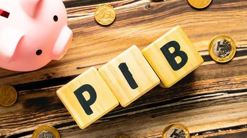 Blocos formando a palavra PIB com moedas e um cofre-porquinho sobre uma mesa