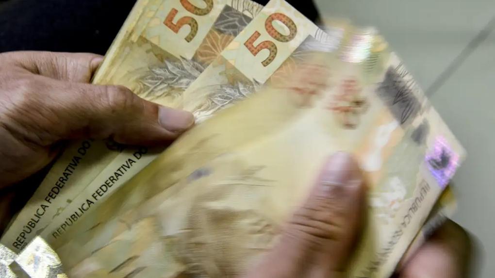 Imagem em close-up mostra pessoa contando cédulas de 50 reais nas mãos. Saiba quando é paga a segunda parcela do décimo terceiro