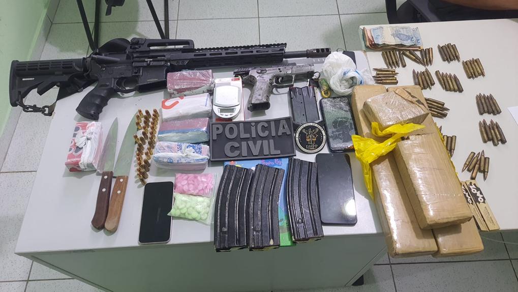 mesa com itens apreendidos: fuzil, pistola, munições, drogas e dinheiros