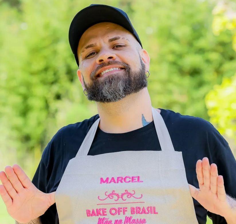 Marcel é um homem branco, barbudo e usa boné preto e camisa da mesma cor. Na foto, ele está de avental do programa Bake Off Brasil