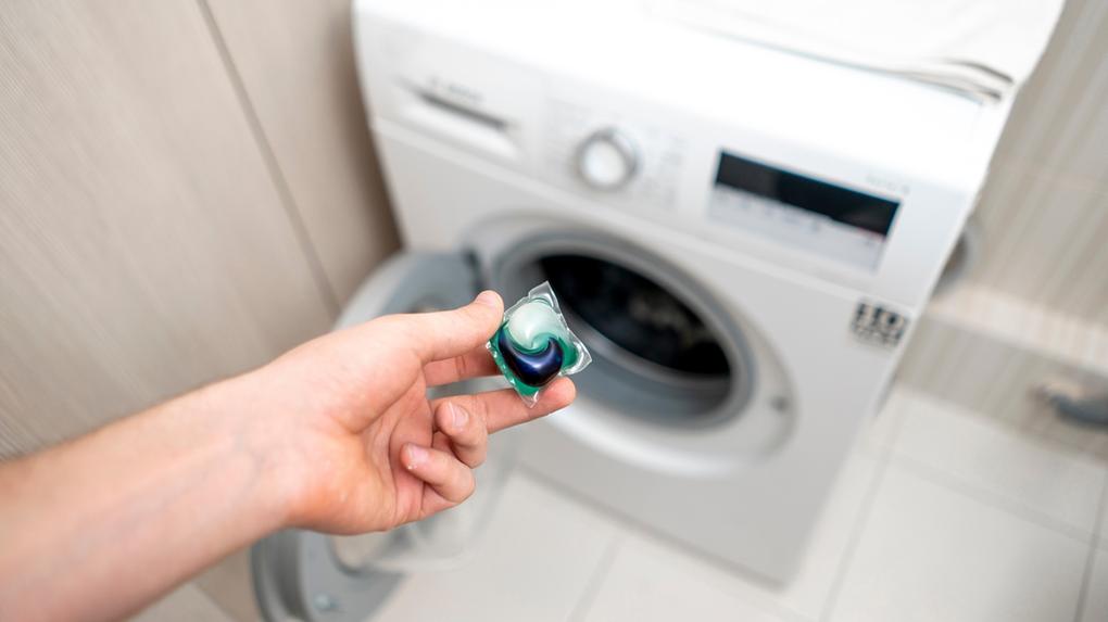 Imagem mostra mão segurando cápsula de detergente para roupas e, ao fundo, uma máquina de lavar roupas. 3 pessoas são internadas após ingerirem detergente para roupas distribuído por político em Taiwan
