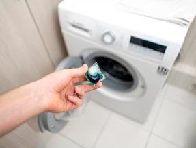 Imagem mostra mão segurando cápsula de detergente para roupas e, ao fundo, uma máquina de lavar roupas. 3 pessoas são internadas após ingerirem detergente para roupas distribuído por político em Taiwan