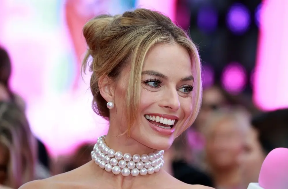 Margot Robbie de cabelo preso sorrindo. Na foto, ela usa um colar de pérolas de três camadas