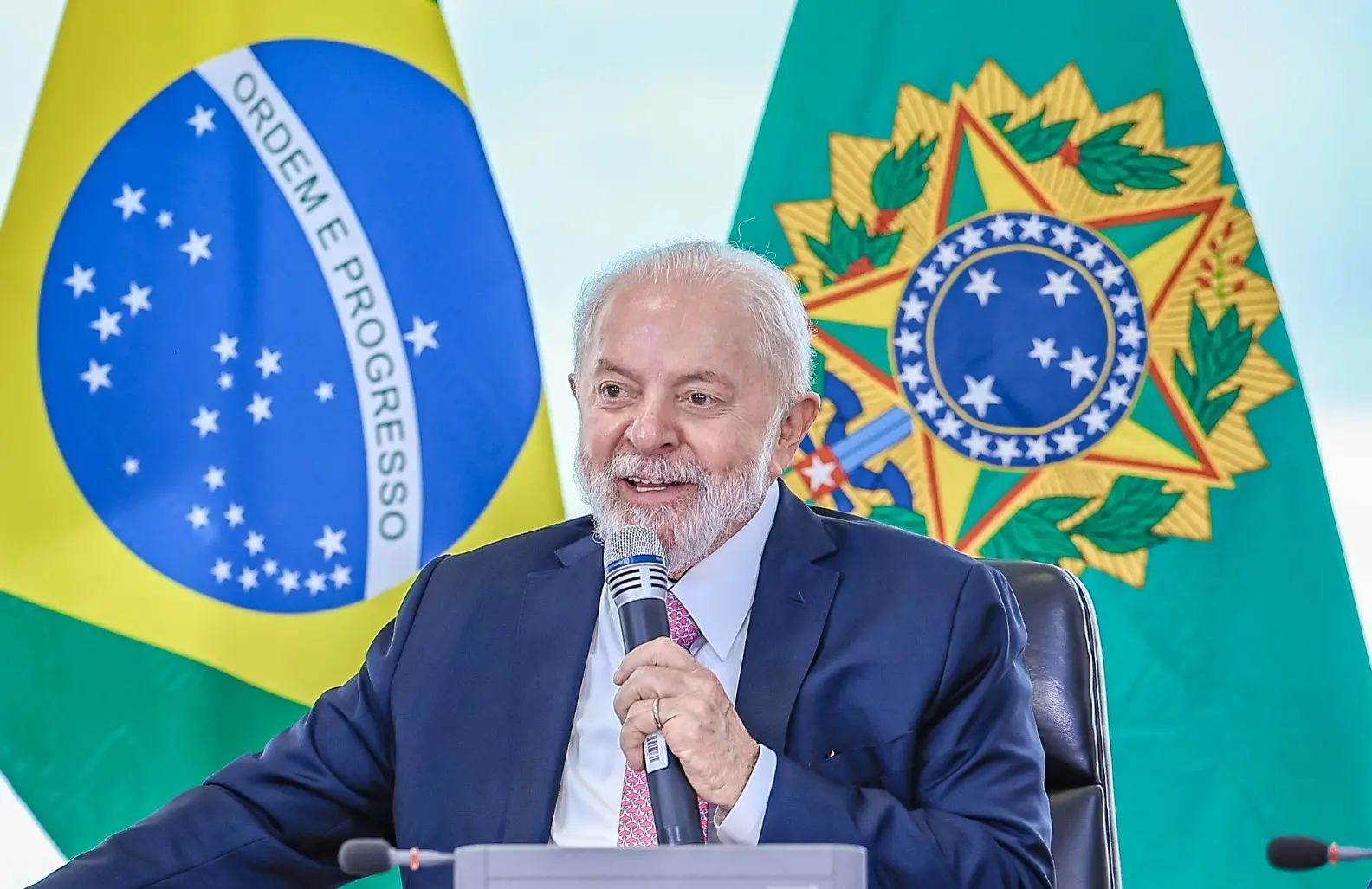 Presidente Lula segura microfone enquanto discursa em frente à bandeira do Brasil