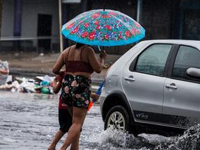 Mulher usando um guarda-chuva segura na mão de uma criança e atravessa uma via alagada, próxima a um carro, em um dia chuvoso em Fortaleza, Ceará. Oito municípios do Ceará seguem com alerta de risco de chuvas intensas até está segunda (29)