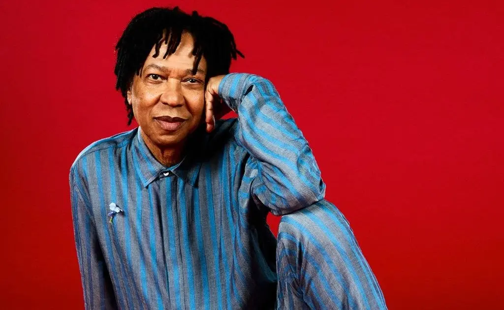 Djavan posa sentado para foto. Ele é um homem negro e está de roupa azul listrada na frente de um fundo vermelho