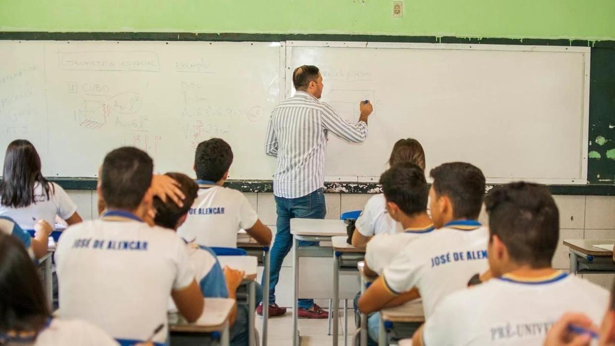 Professor em sala de aula escreve na lousa enquanto é observado por estudantes
