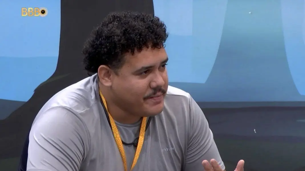 Lucas tem cabelo cacheado e usa bigode. Ele está usando uma camiseta cinza