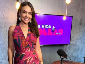 A jornalista Taís Lopes posa diante de uma TV com o anúncio do podcast A Vida é Delas