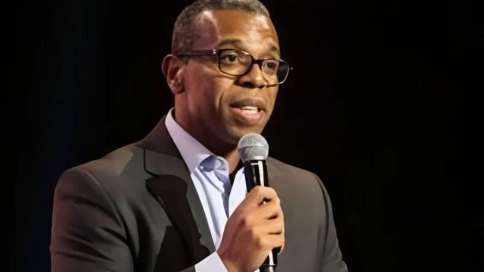 Márcio Santos, na imagem, está de camisa branca e paletó preto, enquanto segura um microfone. Ele é um homem negro, de óculos e de cabelos grisalhos e baixos