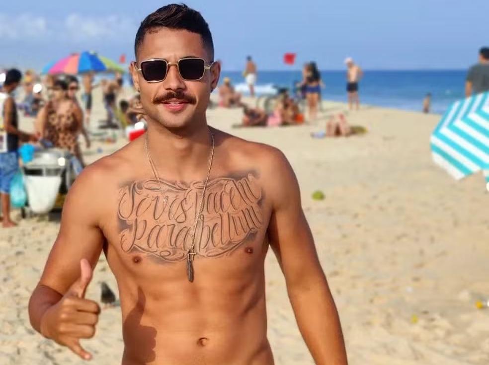 Lucas é um homem jovem, com o peito tatuado e usa bigode. Na foto, ele está sem camisa, na praia, e utiliza óculos escuros