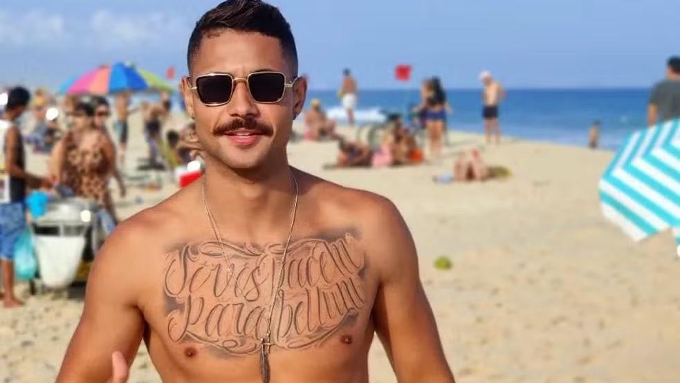 Lucas é um homem jovem, com o peito tatuado e usa bigode. Na foto, ele está sem camisa, na praia, e utiliza óculos escuros