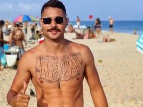 Lucas é um homem jovem, com o peito tatuado e usa bigode. Na foto, ele está sem camisa, na praia, e utiliza óculos escuros