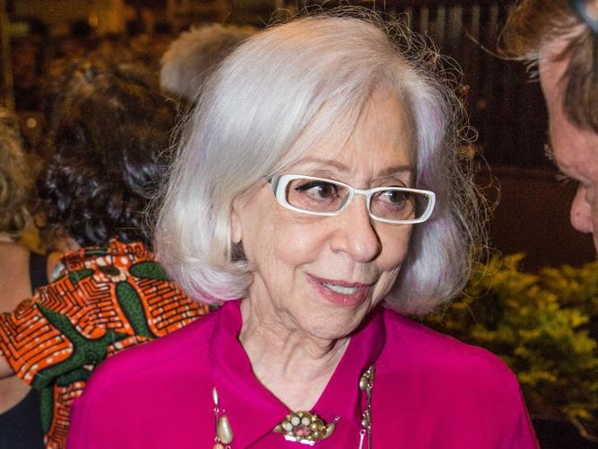 Fernanda Montenegro é uma mulher branca de cabelo médio também branco. Ela usa óculos de armação branca e está vestida com uma camisa rosa de botão