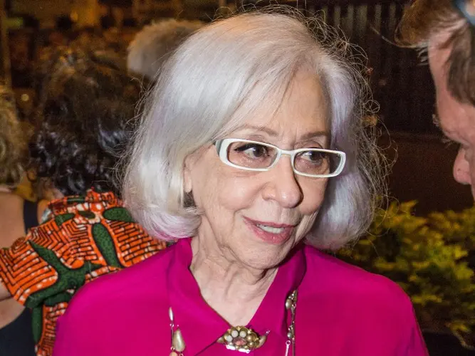 Fernanda Montenegro é uma mulher branca de cabelo médio também branco. Ela usa óculos de armação branca e está vestida com uma camisa rosa de botão