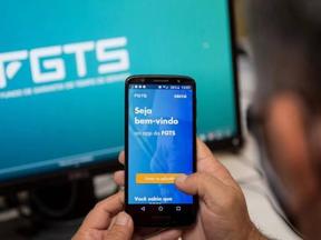 Pessoa com celular abre aplicativo do FGTS em frente a monitor mostrando o site do FGTS