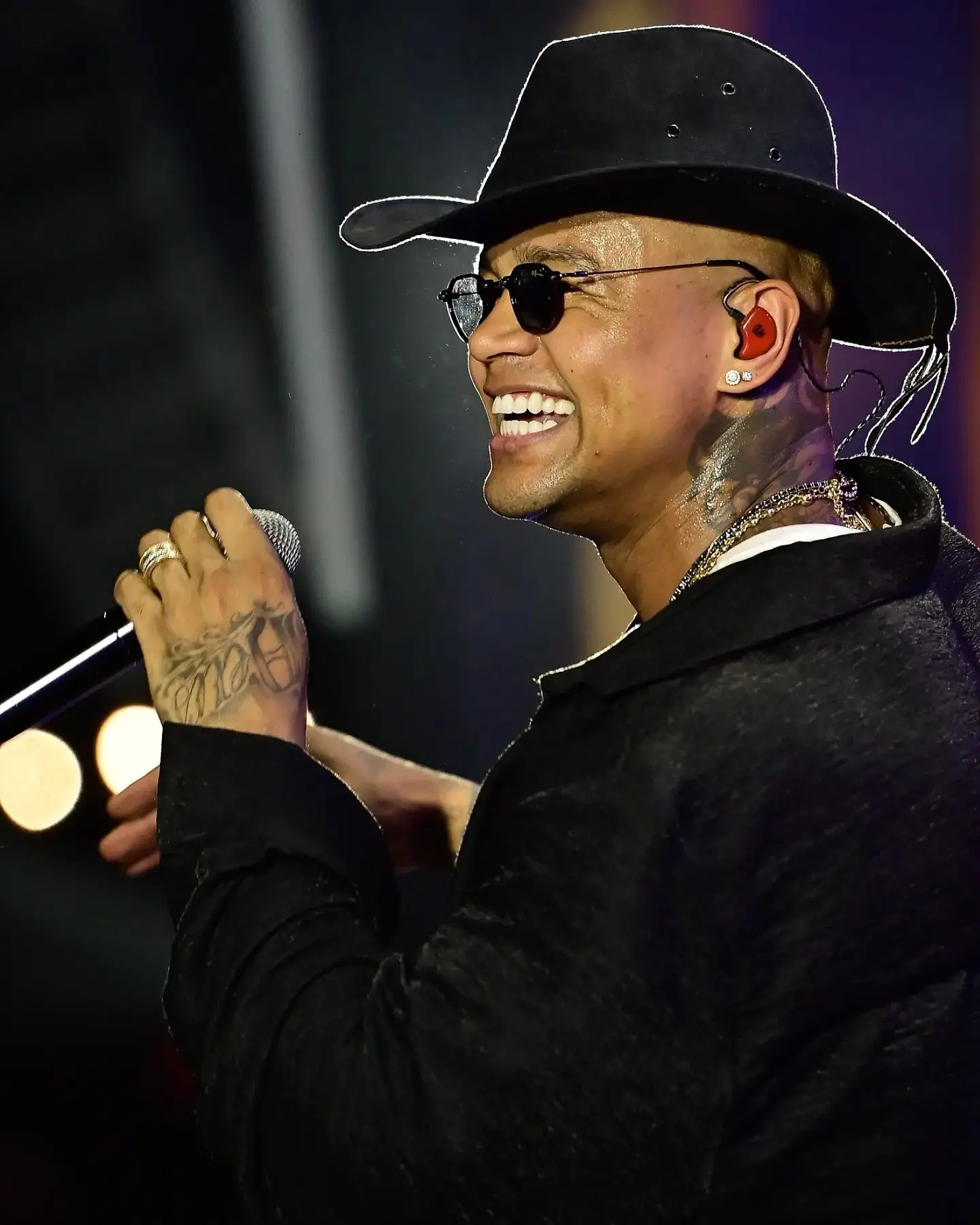 O cantor Leo Santana está de chapéu e óculos escuros, de lado, sorrindo e segurando um microfone