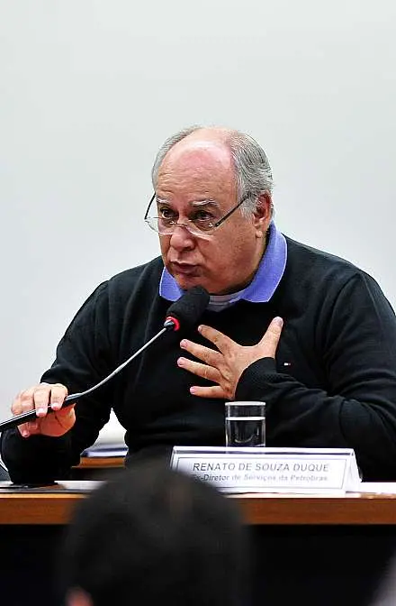 Renato Duque é um homem idoso, branco, calvo, corpulento. Na foto, ele está de camisa preta com uma camisa de gola polo